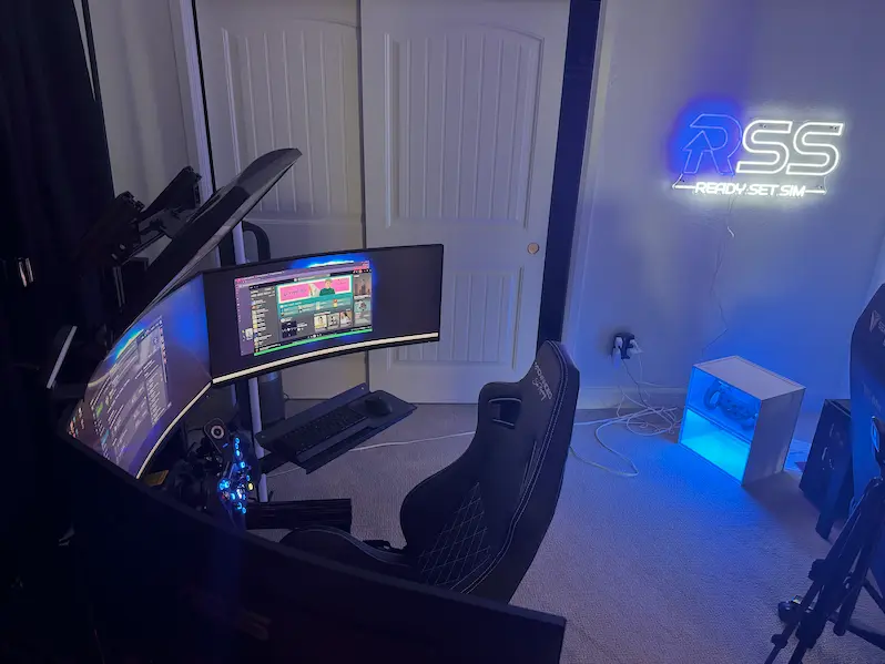 A triple-screen sim rig with the Ready Set Sim neon sign glowing on the wall